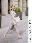 Side view of elegant young feminine businesswoman with long blond hair in stylish outfit, and high heels walking on paved street with laptop in hand and looking away. Self assured female manager with laptop crossing street near building. Стоковое фото, агентство Ingram Publishing / Фотобанк Лори