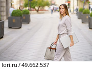 Side view of confident adult female manager in stylish outfit with blond hair looking at camera, while walking on paved street with laptop and bag in hands. Stylish female entrepreneur with netbook strolling on city street. Стоковое фото, агентство Ingram Publishing / Фотобанк Лори