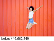 Full body of smiling African American female in casual outfit looking at camera while jumping with outstretched arms and bent knees against red background. Carefree black woman jumping near wall. Стоковое фото, агентство Ingram Publishing / Фотобанк Лори