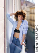 Adult hot female with curly hair touching head and leaning on railing while standing on balcony against blurred background of city street during sunny day. Woman touching curly hair on balcony. Стоковое фото, агентство Ingram Publishing / Фотобанк Лори