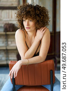 Sensual young female model with Afro hairstyle in black lingerie and jeans sitting on brown chair and touching shoulder while looking away. Woman in bra sitting on chair. Стоковое фото, агентство Ingram Publishing / Фотобанк Лори