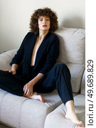 Full body high angle of adult woman with curly dark hair sitting on couch wearing black jacket and pants looking at camera. Stylish curly haired female sitting on sofa wearing black suit. Стоковое фото, агентство Ingram Publishing / Фотобанк Лори