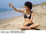 Full body of positive African American female traveler in bikini pouring sand while sitting on coast near sea in tropical resort in sunny weather. Content black woman pouring sand on shore in summer. Стоковое фото, агентство Ingram Publishing / Фотобанк Лори