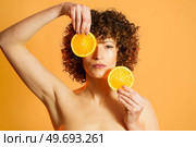 Serious adult female with curly hair covering face with slices of ripe citrus during beauty routine against orange background. Adult woman with pieces of fresh orange. Стоковое фото, агентство Ingram Publishing / Фотобанк Лори