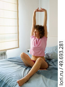 Full body of happy young female in sleepwear sitting on comfortable bed and stretching arms up in morning at home. Relaxed woman stretching arms on bed in bedroom. Стоковое фото, агентство Ingram Publishing / Фотобанк Лори