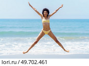 Full body of cheerful young fit African American female tourist, with dark curly hair in yellow bikini doing star jump, and smiling on sandy beach near wavy sea. Excited ethnic woman jumping with raised arms on sandy seashore. Стоковое фото, агентство Ingram Publishing / Фотобанк Лори