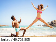 Full body of happy young woman with curly hair jumping on sand with outstretched arms and legs near kneeling African American boyfriend. Cheerful woman jumping on beach near man. Стоковое фото, агентство Ingram Publishing / Фотобанк Лори