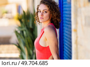 Side view of confident young female in red sundress with curly hair looking at camera, while standing against blue shutters and blurred tropical plants in daylight. Attractive woman in sleeveless dress standing on street. Стоковое фото, агентство Ingram Publishing / Фотобанк Лори