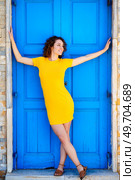 Full body of happy young female in yellow dress with outstretched arms, and crossed legs looking away and smiling while leaning on blue door on street. Cheerful woman leaning on blue door on street. Стоковое фото, агентство Ingram Publishing / Фотобанк Лори