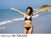 Positive African American female in swimwear with Afro hairstyle looking into distance with outstretched arms while standing on wet coast near sea. Gleeful black woman in bikini standing near sea. Стоковое фото, агентство Ingram Publishing / Фотобанк Лори