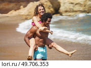 Happy young multiracial couple fooling around on sandy beach near waving ocean while enjoying summer vacation together during sunny weekend. Joyful black man giving piggyback ride to girlfriend near sea. Стоковое фото, агентство Ingram Publishing / Фотобанк Лори