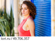 Side view of beautiful young female in red sleeveless sundress standing in front of building with blue shutters and looking away against blurred tropical plants. Young woman in red sundress standing on street. Стоковое фото, агентство Ingram Publishing / Фотобанк Лори