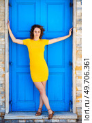 Full body of confident young female in yellow dress and curly hair looking at camera while standing near blue door on street. Calm woman standing in doorway with outstretched arms. Стоковое фото, агентство Ingram Publishing / Фотобанк Лори