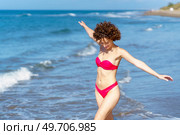Delighted young fit female with dark curly hair, in red bikini smiling and looking down while walking in wavy sea with outstretched arms during summer holidays holidays. Joyful young lady in swimwear walking in sea with outstretched arms. Стоковое фото, агентство Ingram Publishing / Фотобанк Лори