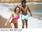 Cheerful multiethnic couple in colorful swimwear running on sandy shore while holding hands having fun together on resort. Joyful multiracial couple fooling around on beach. Стоковое фото, агентство Ingram Publishing / Фотобанк Лори