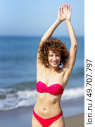 Cheerful young female redhead in pink bikini standing on sandy beach and touching outstretched hands, over head while looking at camera in sunlight against blue seawater and sky. Smiling woman standing with raised hands on beach. Стоковое фото, агентство Ingram Publishing / Фотобанк Лори