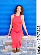Cheerful young female in sleeveless red sundress and sandals looking at camera while relaxing on stone wall against blue shutters in daylight. Smiling woman sitting on stone wall in city street. Стоковое фото, агентство Ingram Publishing / Фотобанк Лори