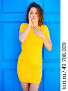 Astonished young female in yellow dress with curly brown hair keeping hand on chest and touching face while looking away against blue wall on street. Amazed woman touching chest and face on street. Стоковое фото, агентство Ingram Publishing / Фотобанк Лори