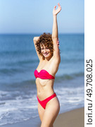 Cheerful young female redhead in pink bikini standing and touching outstretched arm with hand from behind head, while looking at camera in sunlight against blue seawater and sky. Happy woman standing on sandy beach near seawater. Стоковое фото, агентство Ingram Publishing / Фотобанк Лори
