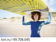 Positive curly haired female in wetsuit carrying SUP board on head smiling and looking at camera while standing on sandy beach on sunny day. Cheerful woman carrying paddleboard on head looking at camera. Стоковое фото, агентство Ingram Publishing / Фотобанк Лори