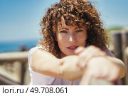 Young curly haired female with freckles and brown wavy hair in white casual clothes stretching arms while leaning on fence against blurred background. Young woman stretching arms on bridge. Стоковое фото, агентство Ingram Publishing / Фотобанк Лори