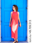 Full body of smiling young female in sleeveless red sundress and flat sandals looking away and standing against blue door in daylight. Happy woman in red dress standing near blue door. Стоковое фото, агентство Ingram Publishing / Фотобанк Лори