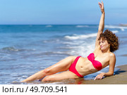 Happy curly woman in bikini with slim body raising arm while lying on beach of ocean. Slim woman in bikini suntanning on sea shore. Стоковое фото, агентство Ingram Publishing / Фотобанк Лори