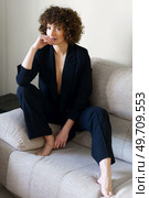 Full body of adult curly haired female wearing black suit sitting on couch touching lips gently and looking at camera. Woman in suit with bare chest sitting on sofa. Стоковое фото, агентство Ingram Publishing / Фотобанк Лори