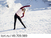 Full body of young female in sportswear and eyeglasses stretching arm overhead to one side while practicing yoga exercise on snowy slope in winter at daylight. Slim woman in sportswear stretching arm sideway in snowy mountains. Стоковое фото, агентство Ingram Publishing / Фотобанк Лори