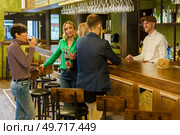 Man ordering some beverages from barkeeper while standing near happy talking women on weekend day in pub. Happy men and women chatting in a beautiful bar. Стоковое фото, агентство Ingram Publishing / Фотобанк Лори
