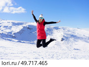 Happy young female in warm clothes cap polarized sunglasses looking at camera, while jumping in air with bent legs and stretched arms in daylight against snowy mountains and blue sky. Carefree young woman jumping with raised arms on snow terrain. Стоковое фото, агентство Ingram Publishing / Фотобанк Лори