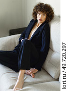 Full body high angle of adult female with curly dark hair sitting on couch wearing black jacket and pants looking at camera. Stylish curly haired woman sitting on sofa wearing black suit. Стоковое фото, агентство Ingram Publishing / Фотобанк Лори