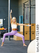 Full body of young sportive blonde female in violet leggings, and white top performing Utkata Konasana with one arm raised and another outstretched while practicing yoga in fitness center. Fit sportswoman doing variation of Goddess pose in gym. Стоковое фото, агентство Ingram Publishing / Фотобанк Лори