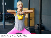 Adult fit female athlete in sportswear and smartwatch, looking away while standing with legs apart and doing exercise with kettlebell during intense workout in blurred gym in daylight. Strong woman doing exercises with kettlebell in gym. Стоковое фото, агентство Ingram Publishing / Фотобанк Лори
