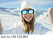 Positive young female in warm clothes beanie cap, and polarized sunglasses smiling and looking at camera while taking selfie and standing on snowy mountain slope on sunny day in winter. Cheerful woman in sunglasses taking selfie in snowy mountains. Стоковое фото, агентство Ingram Publishing / Фотобанк Лори