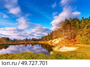 Autumn river landscape. Sunset colorful forest on riverbank. Spruce trees on shore. Beautiful fall calm nature scenery. White clouds on November deep blue sky. Wood on hill. Europe, Belarus. Стоковое фото, агентство Ingram Publishing / Фотобанк Лори