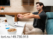 Side view of young male freelancer in casual clothes sitting near laptop and notebook while stretching arms at home. Happy young man stretching arms near netbook at home. Стоковое фото, агентство Ingram Publishing / Фотобанк Лори