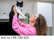 Side view of cheerful young female with curly brown hair, lifting up black and white furred cat looking at camera while playing and bonding at home. Happy young woman spending time playing with cute cat. Стоковое фото, агентство Ingram Publishing / Фотобанк Лори