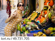 Side view of young stylish female model in trendy armless gown and eyeglasses standing on street and leaning towards stall with assorted fruits on street. Stylish woman leaning toward stall with assorted fruits on street. Стоковое фото, агентство Ingram Publishing / Фотобанк Лори