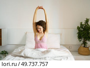Happy adult female with curly hair and eyes closed, while sitting on cozy bed in daylight and stretching reverse clasped hands above head with interlocked fingers in bedroom. Adult woman sitting on bed in sleepwear. Стоковое фото, агентство Ingram Publishing / Фотобанк Лори