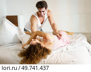 Smiling adult couple in nightwear looking at each other, while male kneeling down with hand on curly haired female waist lying on bed with pillow in daylight in cozy bedroom. Happy couple in sleepwear on bed in daylight. Стоковое фото, агентство Ingram Publishing / Фотобанк Лори
