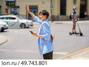 Side view of serious female in formal suit with smartphone at hand standing on city street and catching cab on street. Confident woman making stop sign to taxi on city street. Стоковое фото, агентство Ingram Publishing / Фотобанк Лори