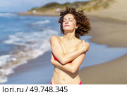 Gorgeous young female redhead relaxing with hands crossed, and touching bare shoulder while facing sun with eyes closed in daylight and standing in beach against blurred sea and sky. Young woman standing on sandy beach on sunny day. Стоковое фото, агентство Ingram Publishing / Фотобанк Лори