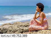 Pensive young African American female tourist, in maxi dress with curly hair, leaning on hand and looking at waving sea while sitting on rough stone on summer day on beach. Thoughtful black woman admiring sea. Стоковое фото, агентство Ingram Publishing / Фотобанк Лори