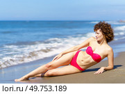 Full length of content curly haired female in bikini lying on arm on sandy beach and looking away in sunlight. Young calm female lying on ocean shore. Стоковое фото, агентство Ingram Publishing / Фотобанк Лори