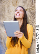 A cheerful young woman wearing a vibrant yellow sweater beams with a bright smile while she holds a tablet, savoring a delightful moment that blends joy with technology against a rustic wall backdrop. A Happy Young Woman Enjoying Some Engaging Digital Content While Outdoors in the Sun. Стоковое фото, агентство Ingram Publishing / Фотобанк Лори