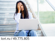 A young woman sits on stairs, overwhelmed and stressed, fixated on her laptop screen. Her expressions reveal frustration and anxiety, highlighting the pressures of modern life and studying. A Stressed Woman Working on Her Laptop While Sitting on the Stairs She Feels Overwhelmed by Work. Стоковое фото, агентство Ingram Publishing / Фотобанк Лори