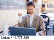 A successful businesswoman in a stylish blazer celebrates her achievement while working on her laptop outdoors, her cheerful expression reflecting professional success and positive energy. Joyful Businesswoman Celebrating Her Success at an Outdoor Workspace with Enthusiasm. Стоковое фото, агентство Ingram Publishing / Фотобанк Лори