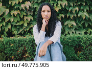A young woman with beautifully curly hair sits in a thoughtful pose in front of lush green foliage, completely embodying a sense of serenity and deep reflection, truly capturing a peaceful moment. A contemplative young woman sitting quietly amidst the vibrant greenery surrounding her. Стоковое фото, агентство Ingram Publishing / Фотобанк Лори
