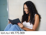 A smiling young woman with beautiful curly hair is completely engrossed in a fascinating and captivating book, dressed casually in a serene indoor setting which radiates joy and relaxation. A Young Woman is Found Enjoying a Book in a Cozy Indoor Setting, Immersed in Reading Delightfully. Стоковое фото, агентство Ingram Publishing / Фотобанк Лори